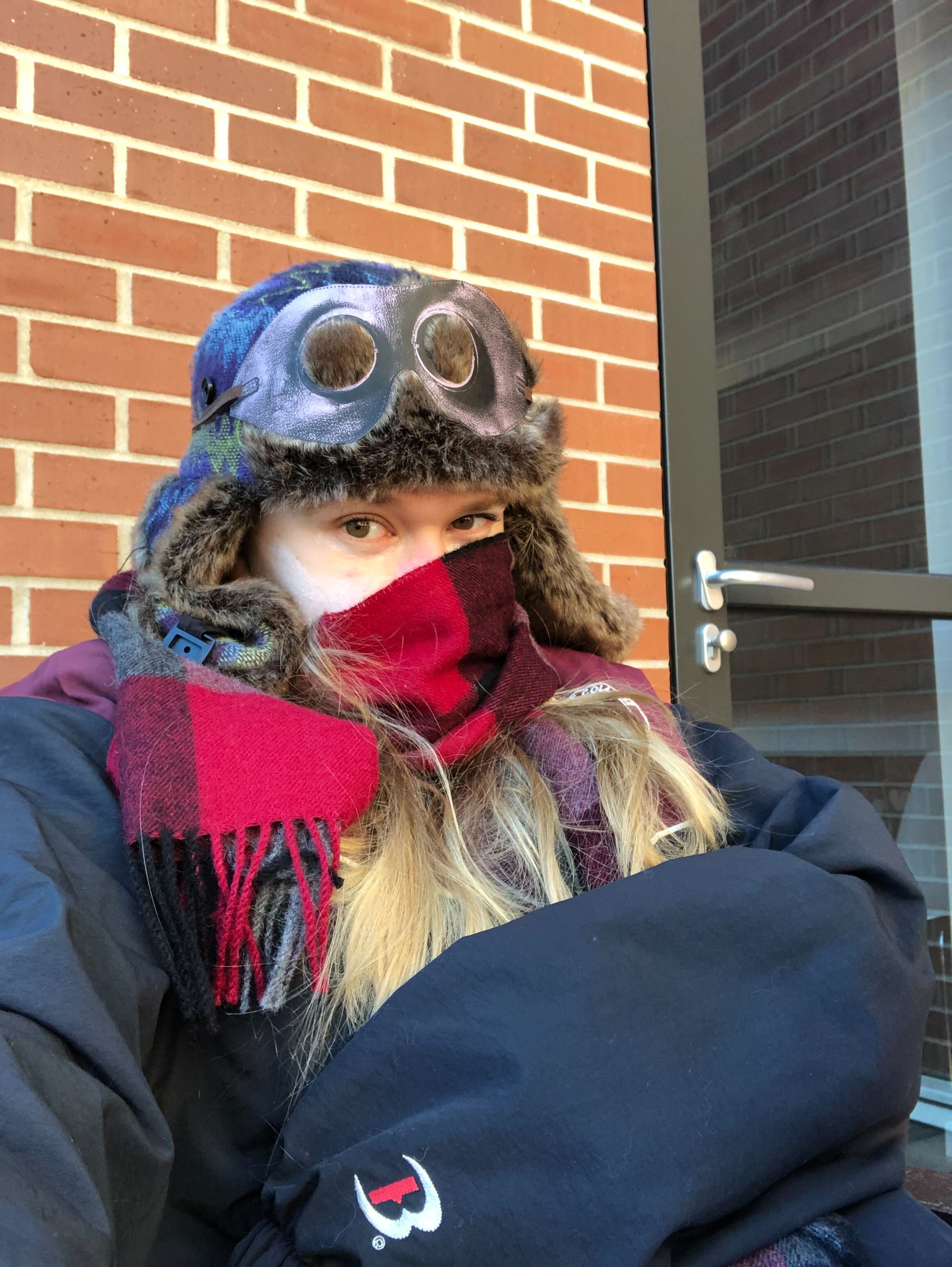 Ellory sits outside on the patio with a brick wall behind her. She is bundled up in a coat with a scarf around her face and a large winter hat on her head.