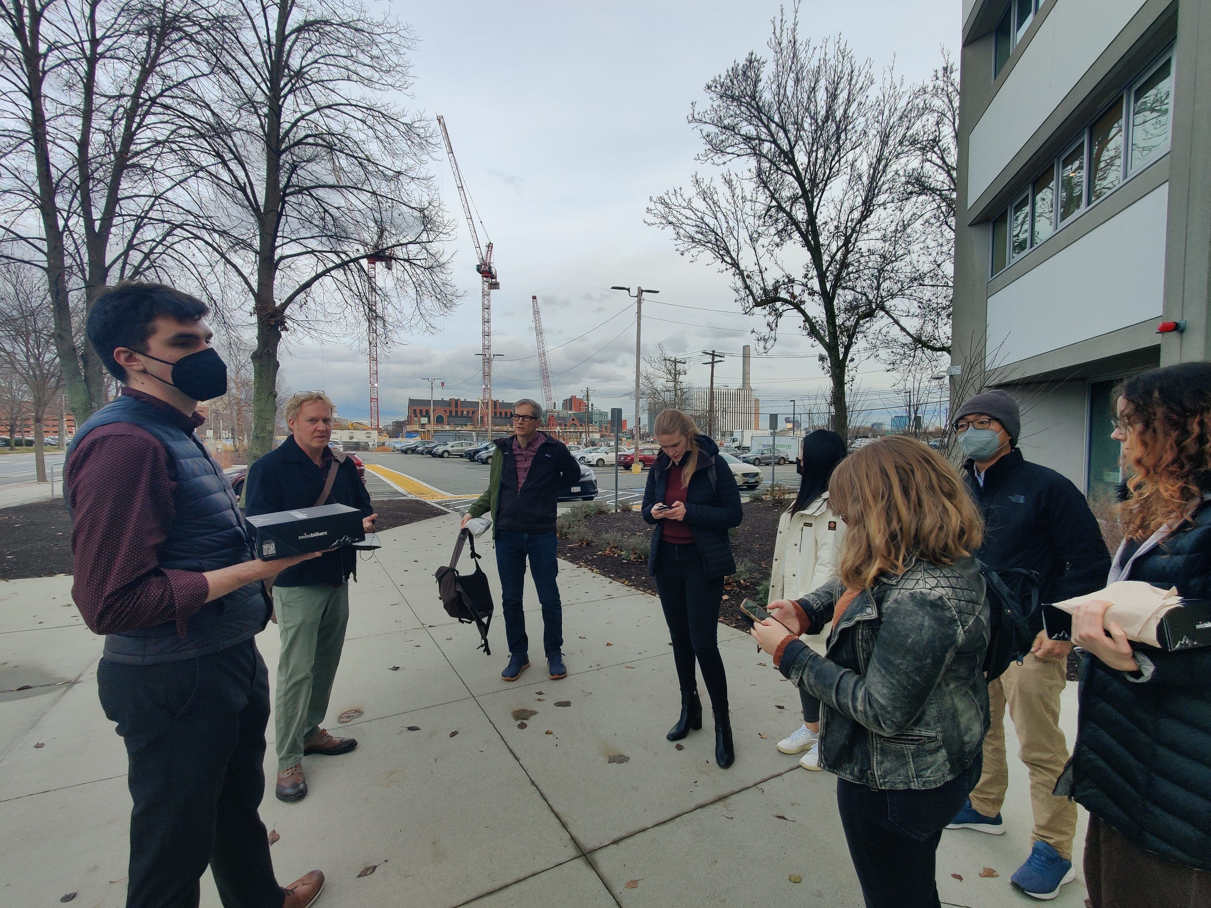 A group of people standing in a loose circle outside a building, looking around or at their phones.