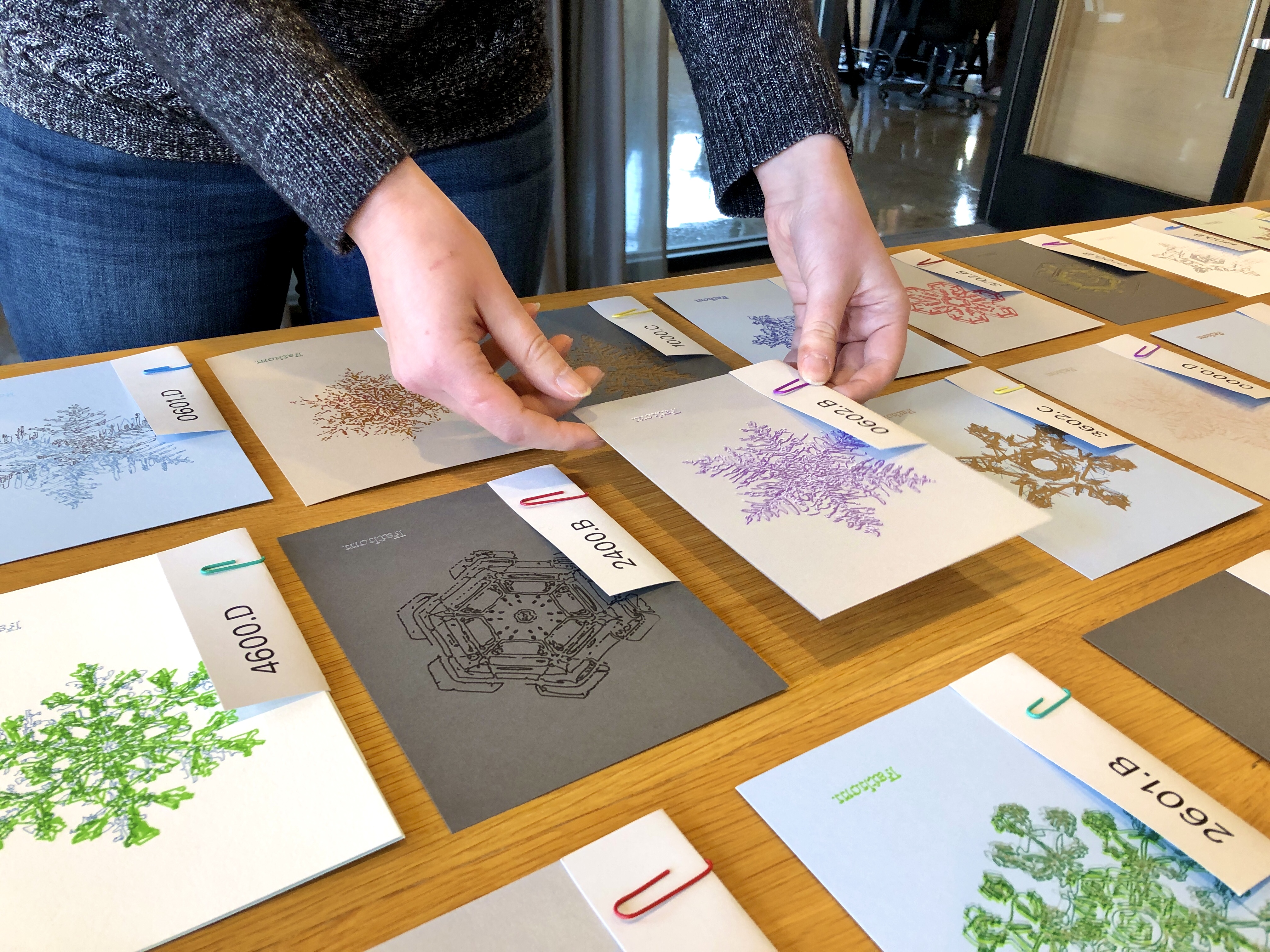 Snowflake holiday cards laid out densely on a big table, each with a slip of paper labeling the ID of its unique design