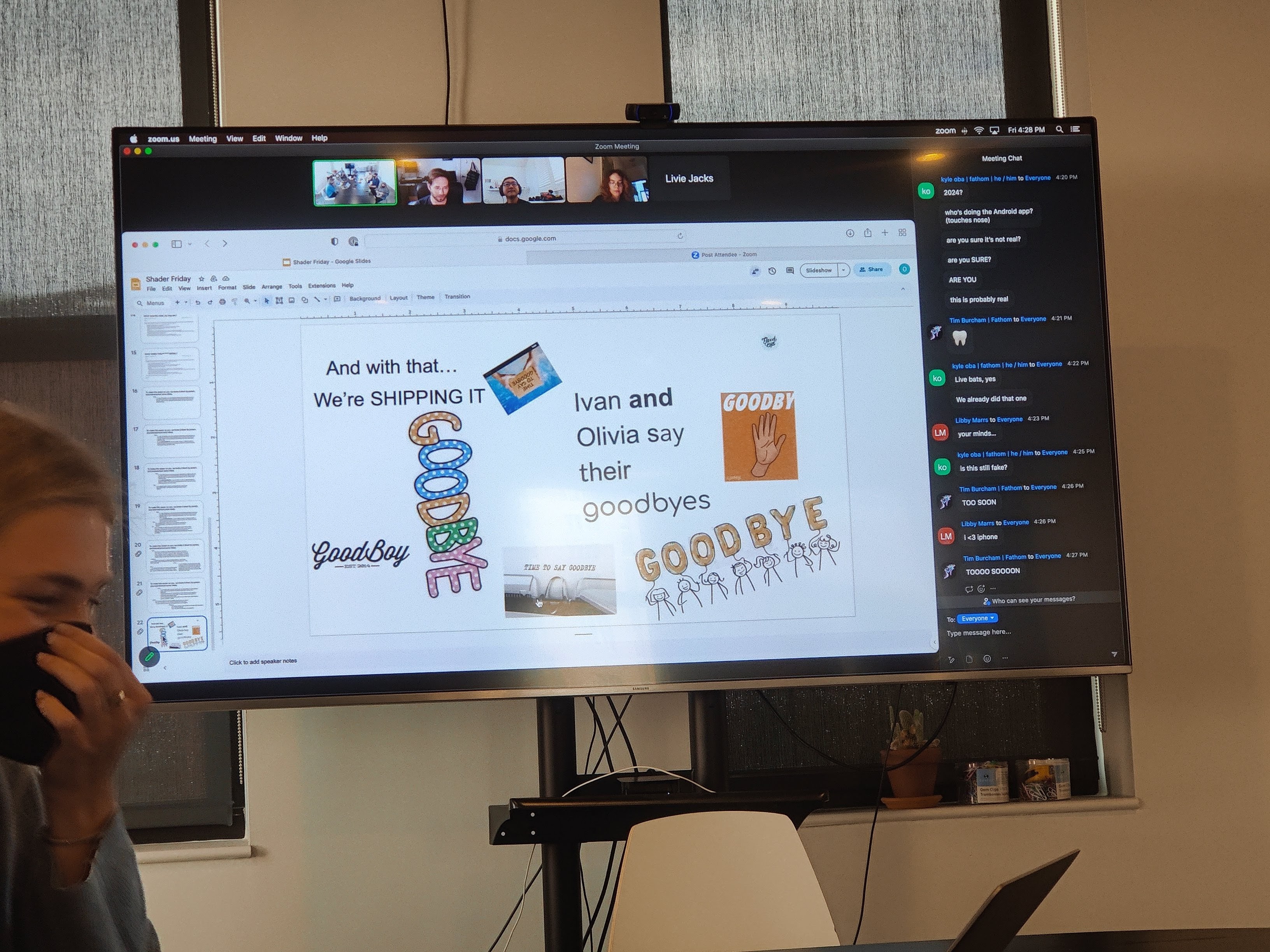 A monitor showing a Zoom call, currently screen sharing to a slideshow that reads, “And with that… We’re SHIPPING IT. Ivan and Olivia say their goodbyes.” The blocks of text are surrounded by cheesy clip art (mis-)representations of “goodbye”, including the logo of GoodBoy, the clothing brand. Just visible at the edge of the photo is Livie, who is holding a hand to her mask, mid-laugh.