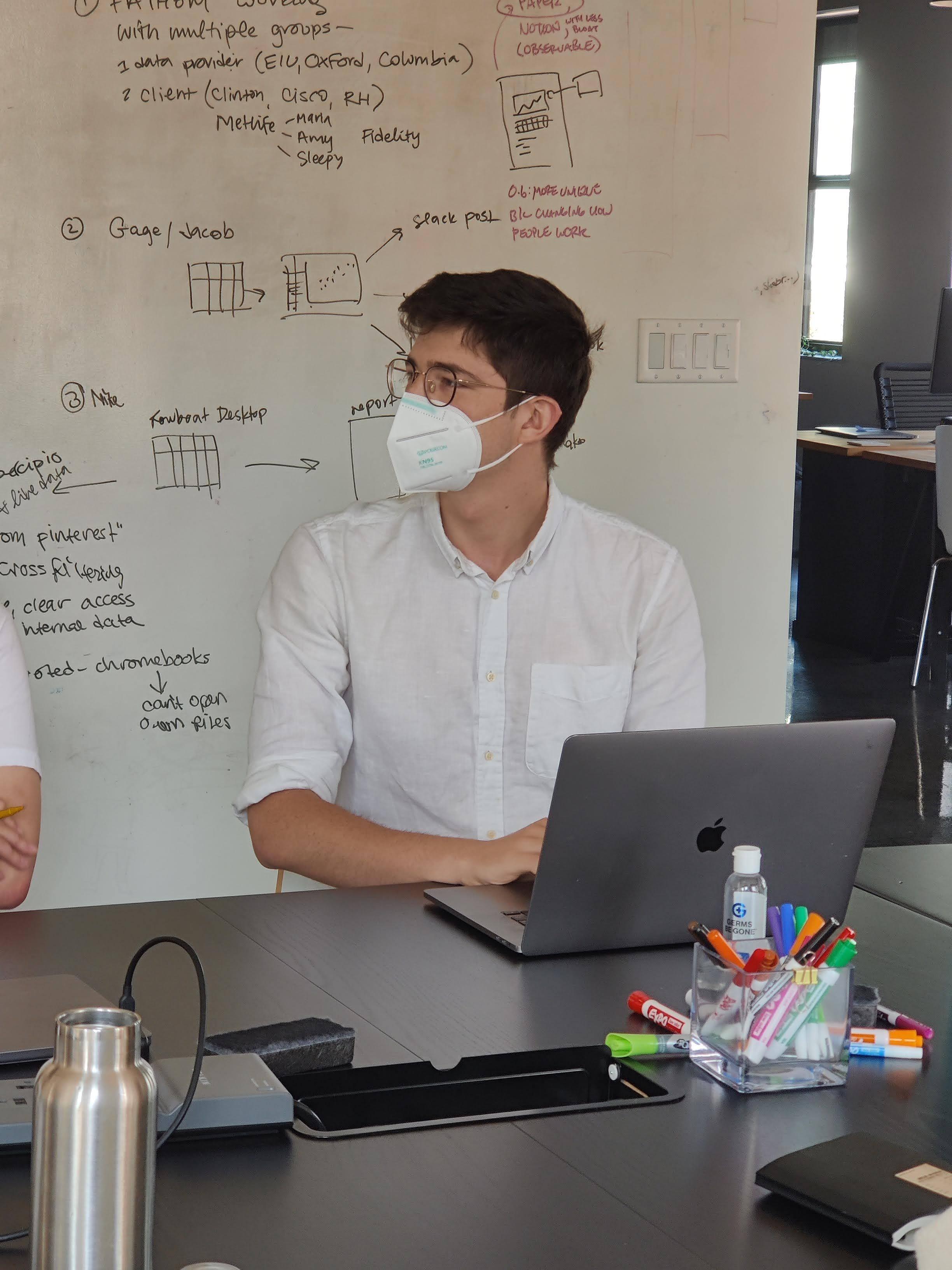 Ivan sits at the conference table with his laptop, wearing a mask and glasses and looking towards the front of the room. Behind him is a whiteboard wall with scribbled notes.