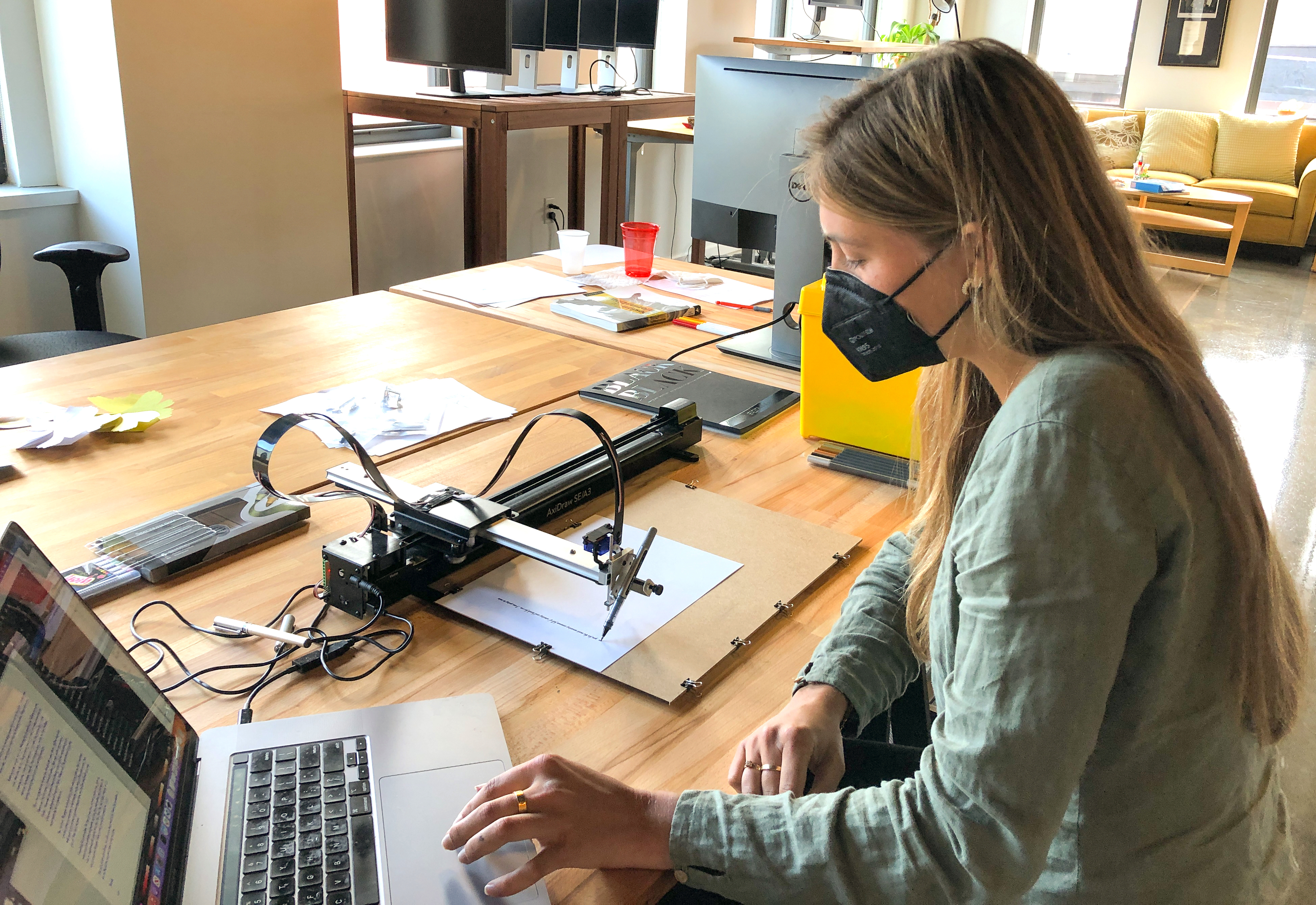 Livie sits at the AxiDraw station with her laptop, watching the plotter draw a line of text.